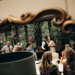 Pocałunek pary młodej uchwycony w ozdobnym lustrze podczas przyjęcia weselnego w oranżerii, z gośćmi w tle. Ciepłe światło.