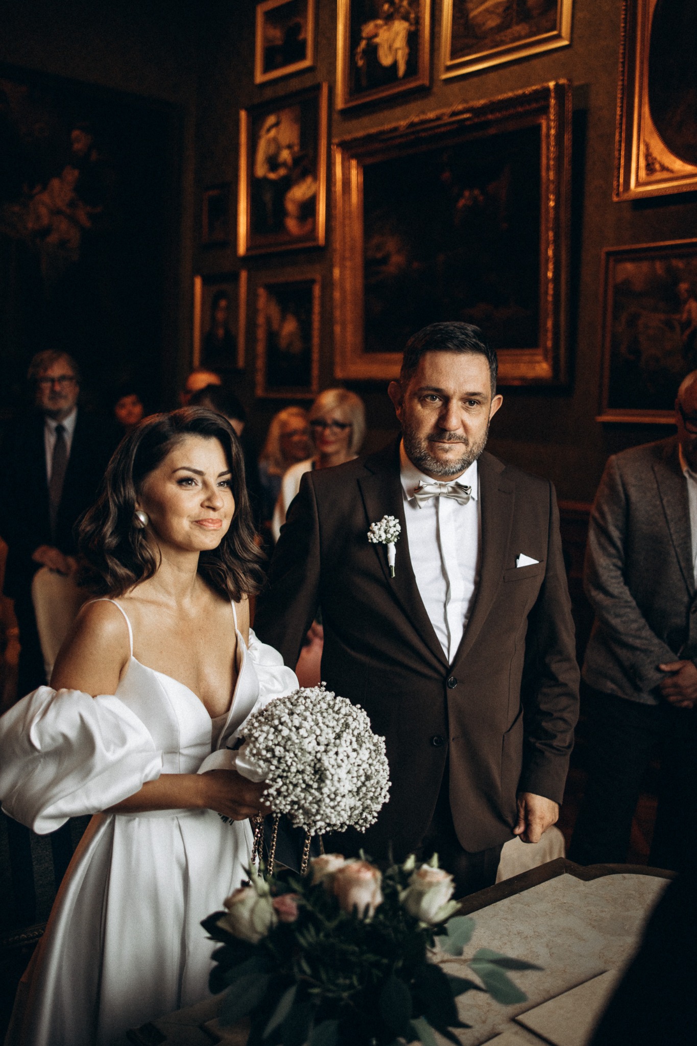 Elegancka para młoda podczas ceremonii ślubnej w bogato zdobionym wnętrzu. Panna młoda w białej sukni, pan młody w brązowym garniturze. W tle obrazy w złotych ramach.