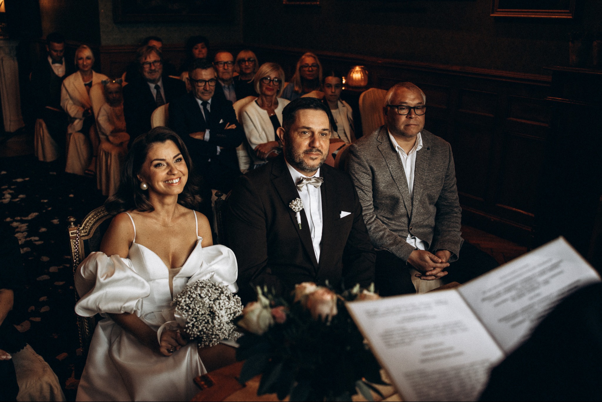 Uśmiechnięta panna młoda z bukietem i pan młody w muszce podczas ceremonii ślubnej, na tle gości w ciemnym wnętrzu.