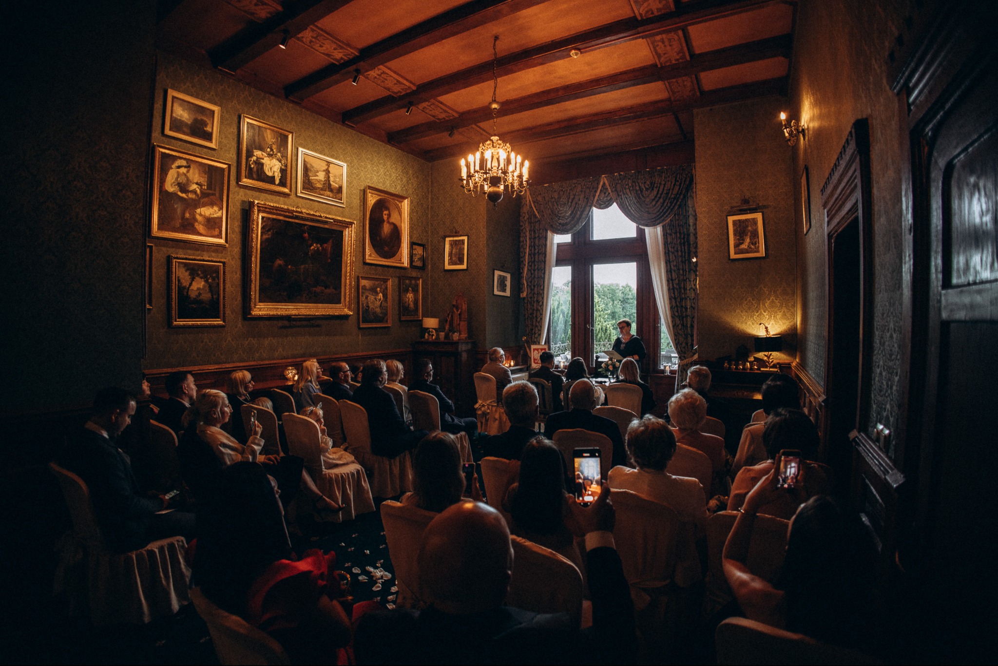 Eleganckie wnętrze z publicznością podczas ceremonii. Ściany zdobione obrazami, żyrandol, osoba przemawiająca przy oknie. Goście robią zdjęcia telefonami.