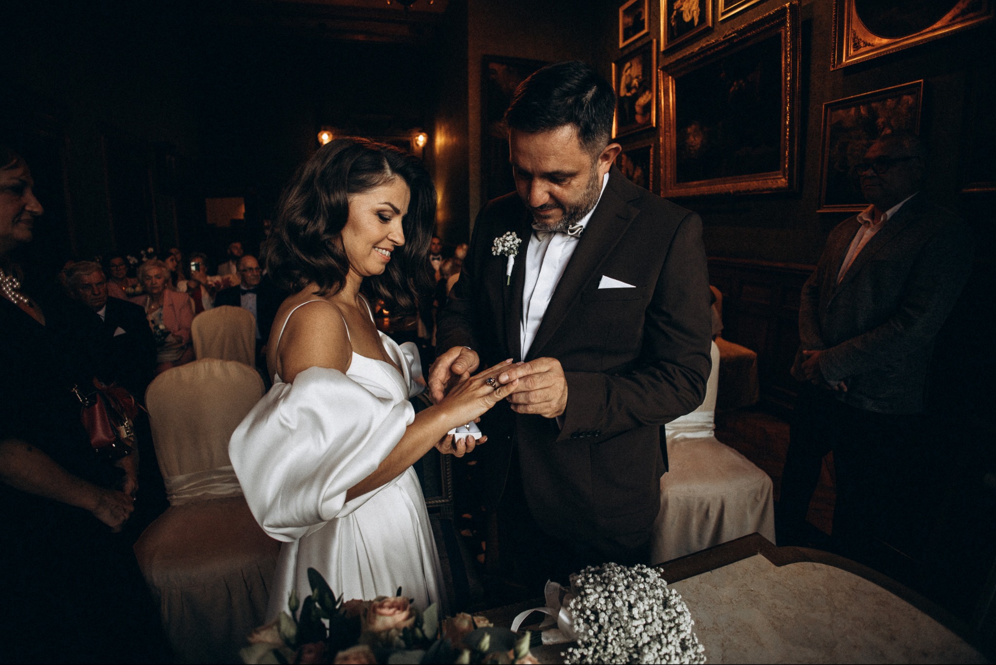 Para wymienia się obrączkami podczas ceremonii ślubnej w eleganckim wnętrzu. Panna młoda w białej sukni, pan młody w brązowym garniturze. W tle goście i obrazy.