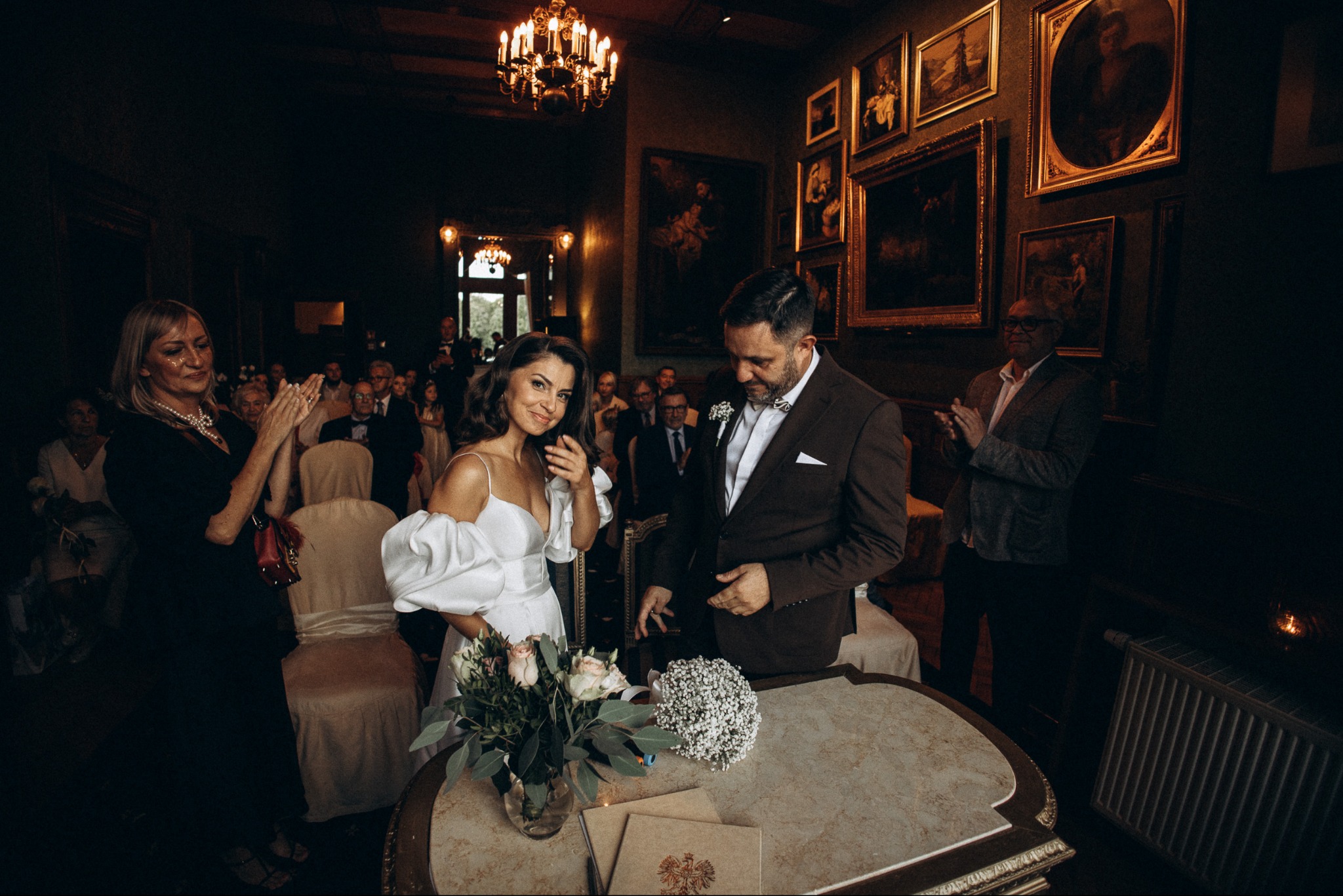 Para młoda w eleganckim wnętrzu, otoczona gośćmi podczas ceremonii zaślubin. W tle obrazy w złotych ramach i żyrandol.