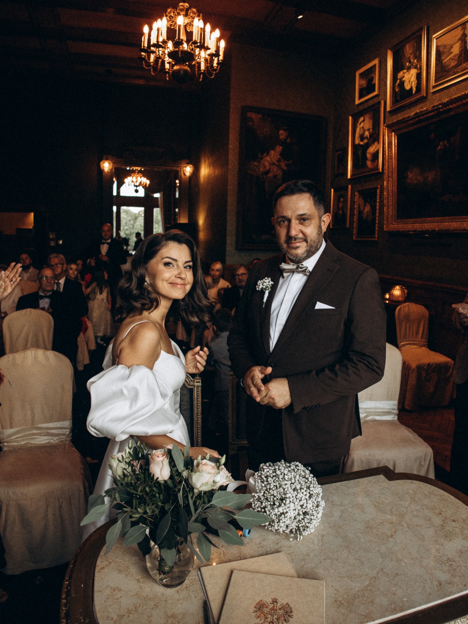 Elegancka para młoda w zabytkowym wnętrzu podczas ceremonii ślubnej. Panna młoda w białej sukni, pan młody w brązowym garniturze z muchą. W tle goście i obrazy.