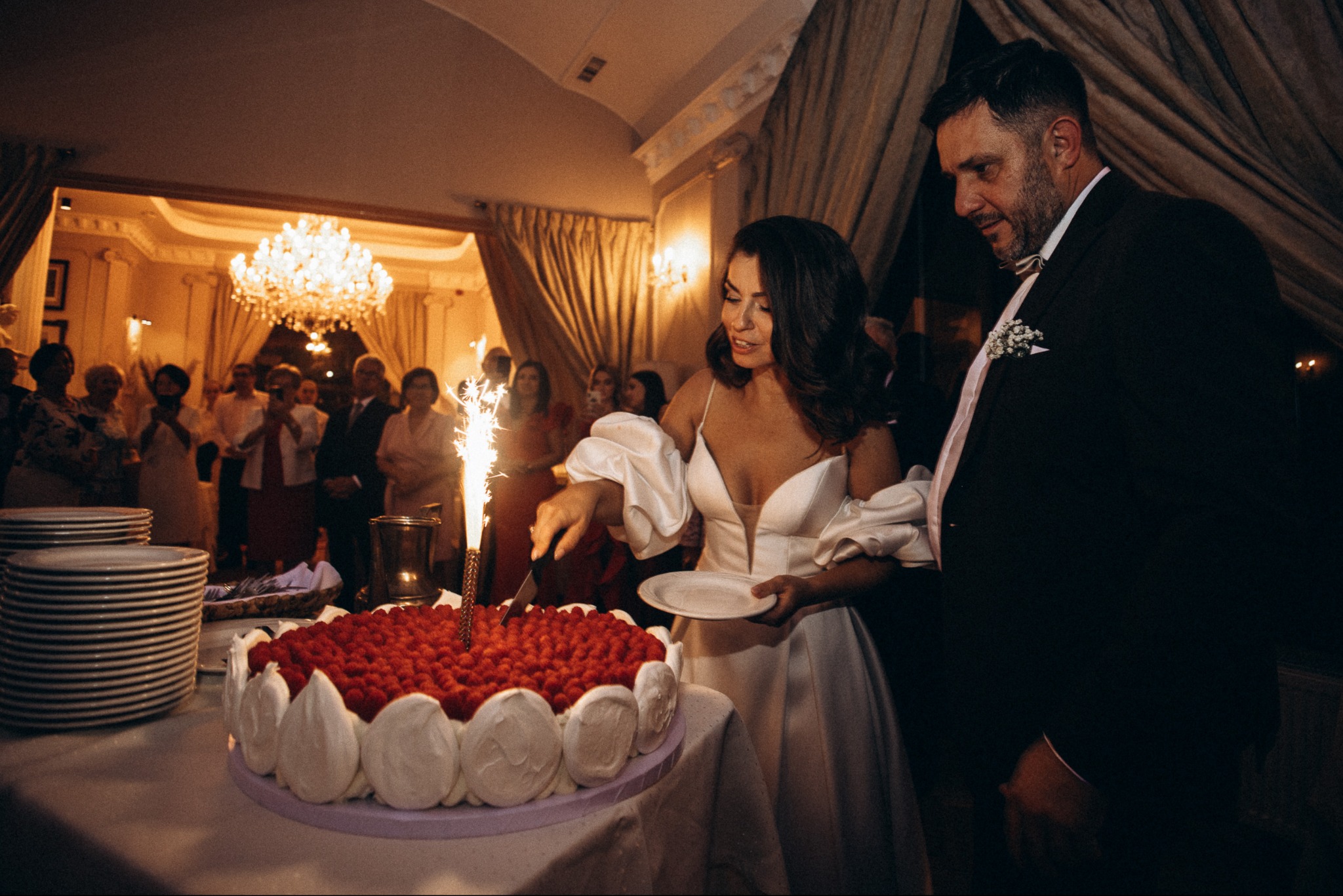 Elegancka para kroi tort weselny z płonącą świeczką na tle gości i kryształowego żyrandola. Ujęcie z bliska, ciepłe oświetlenie, uroczysta atmosfera.