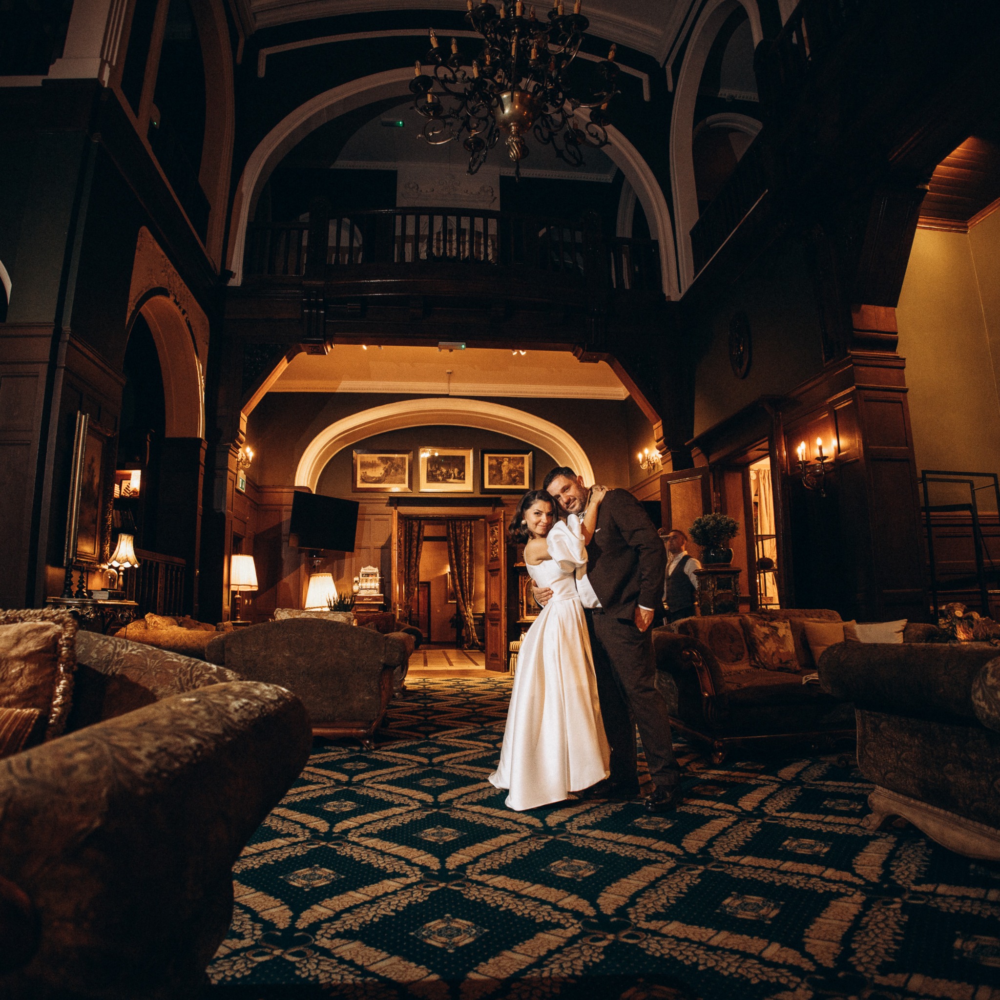 Uśmiechnięta para młoda w eleganckim holu hotelowym z żyrandolem i antycznymi meblami. Ona w białej sukni, on w brązowym garniturze. Romantyczna sesja ślubna.