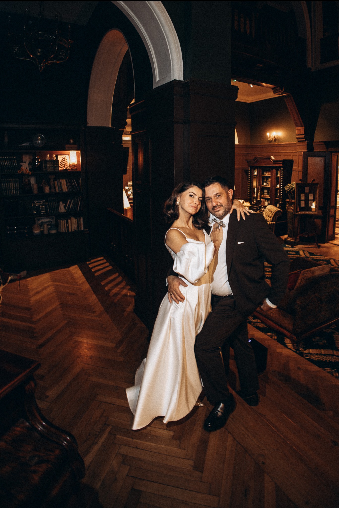 Elegancka para w objęciach na tle wnętrza w stylu retro. Kobieta w białej sukni, mężczyzna w ciemnym garniturze, parkiet w jodełkę, biblioteczka w tle.