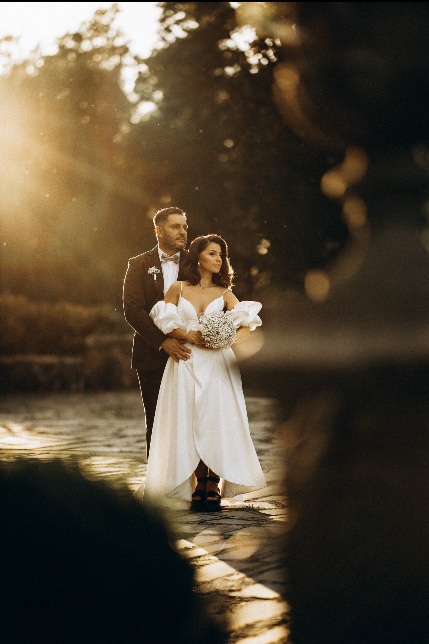 Para młoda w objęciach, ona w sukni z bufiastymi rękawami, on w garniturze z muszką, złote światło słoneczne, naturalna sceneria, romantyczny nastrój.