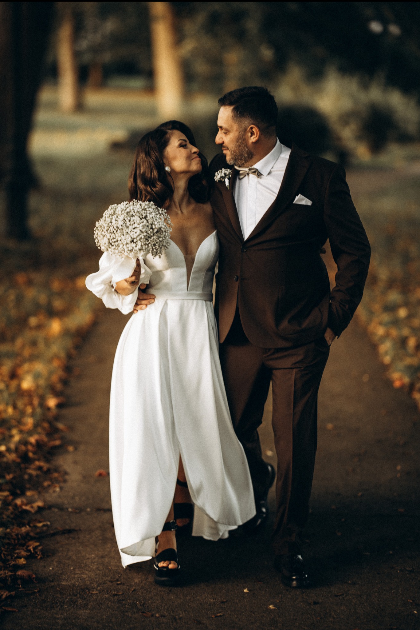 Para młoda w jesiennej scenerii: Panna Młoda w białej sukni z bukietem, Pan Młody w brązowym garniturze, spacerują po ścieżce, patrząc na siebie z czułością.