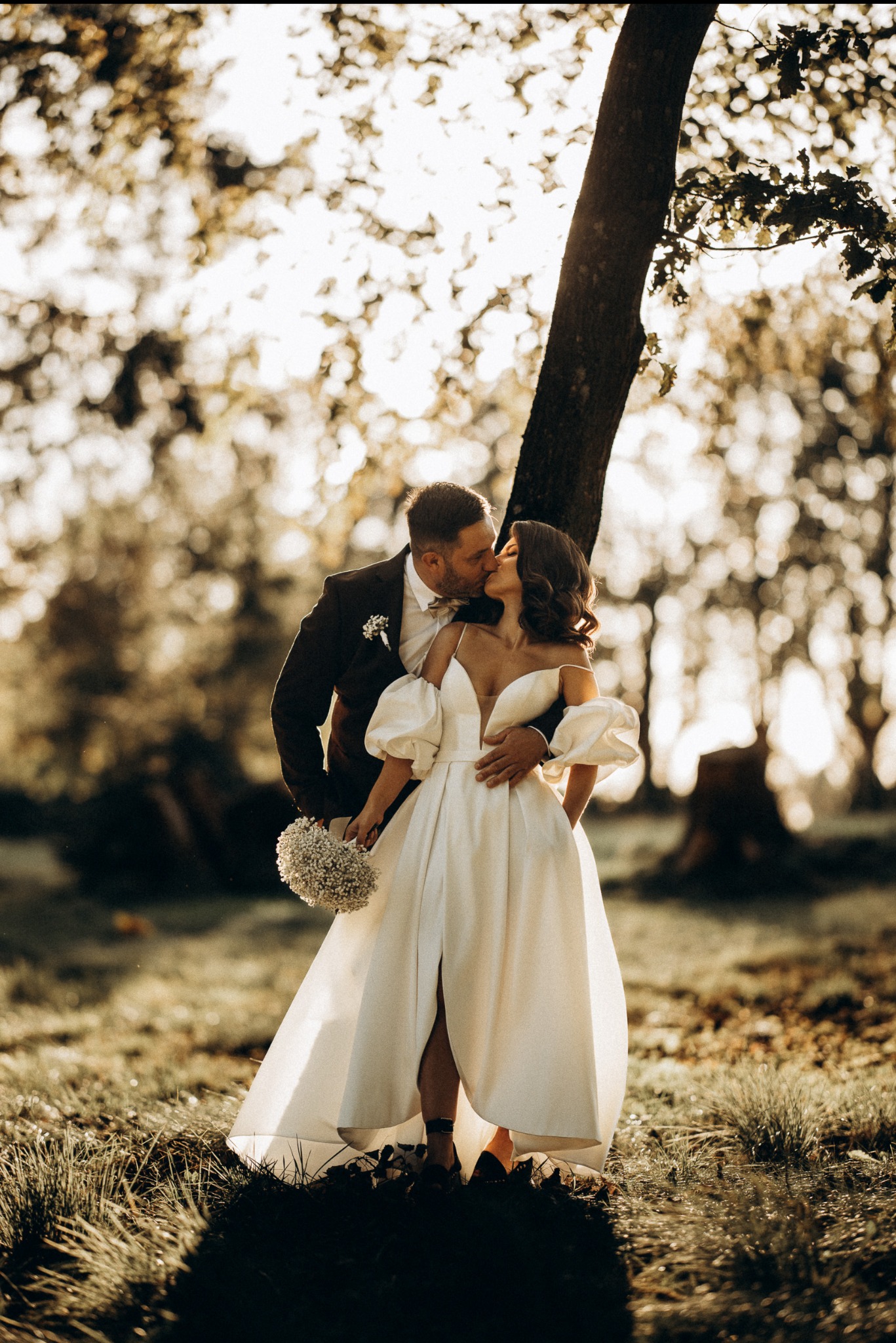 Para młoda w jesiennej scenerii: panna młoda w sukni z bufiastymi rękawami i rozcięciem, pan młody w ciemnym garniturze, czuły pocałunek pod drzewem w złocistym świetle.