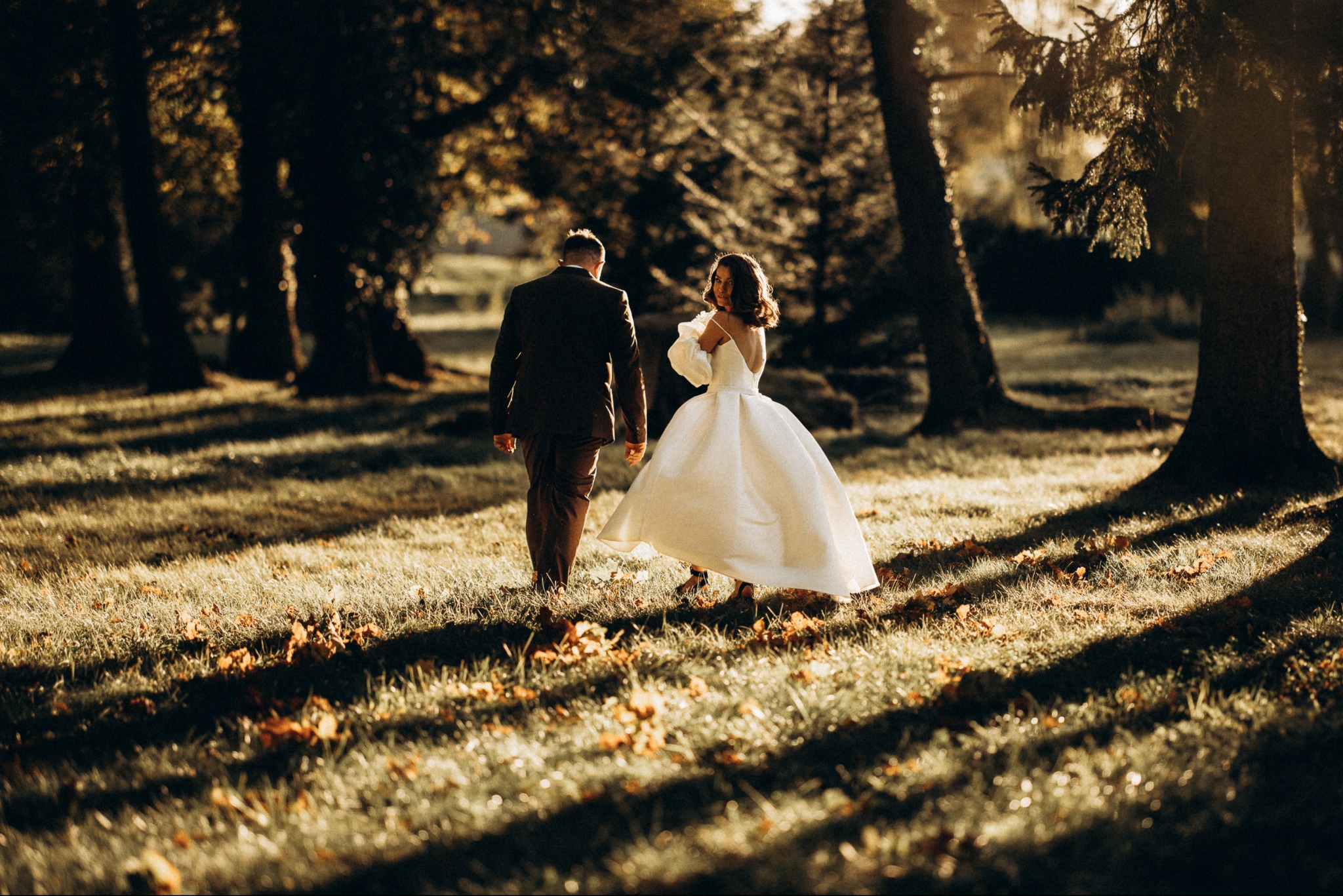 Para w sukni ślubnej i garniturze spaceruje po parku w promieniach zachodzącego słońca. Panna młoda spogląda przez ramię, uśmiechając się.