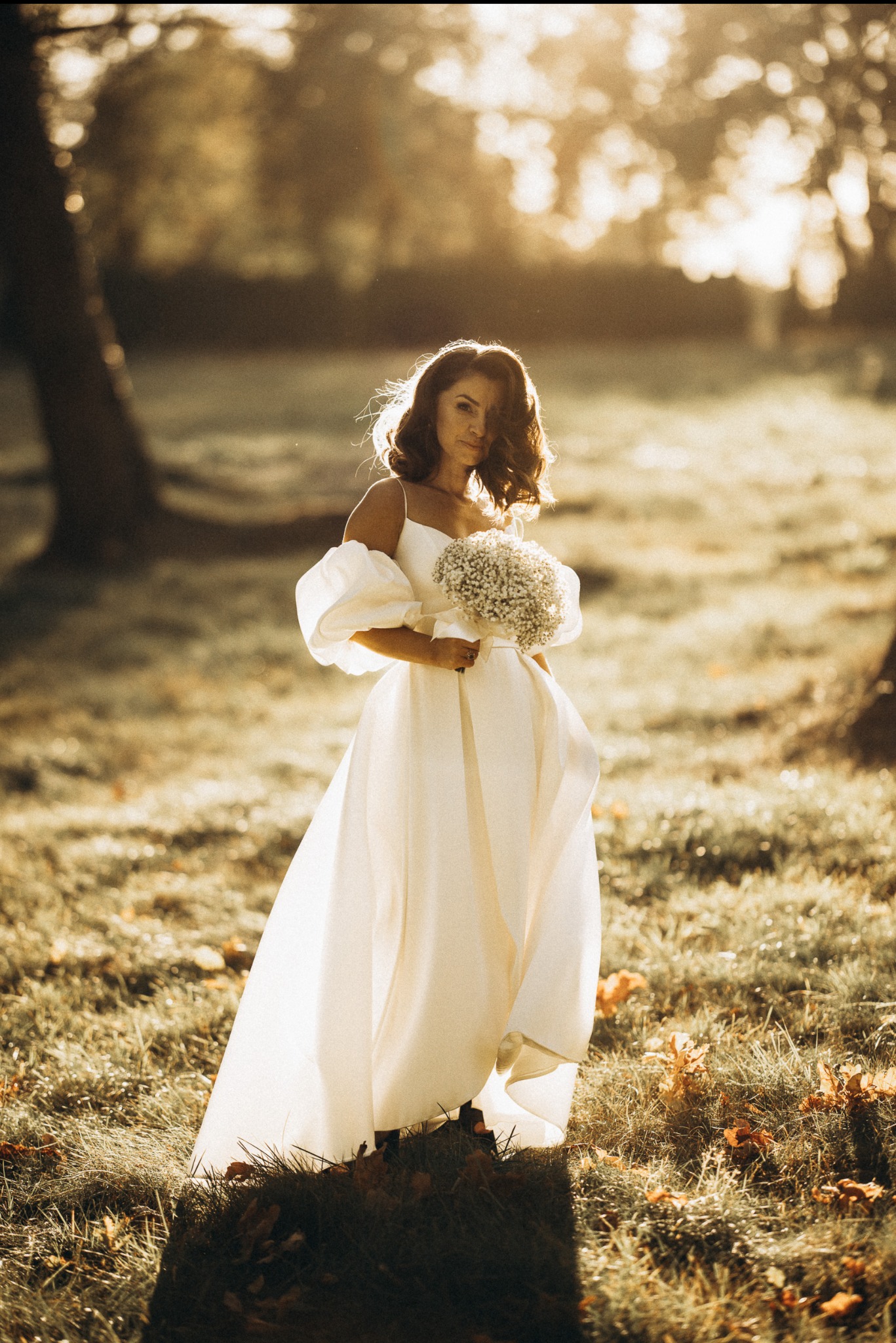 Panna młoda w białej sukni z bukietem polnych kwiatów na łące w promieniach zachodzącego słońca. Romantyczna sceneria z drzewami w tle. Delikatne światło podkreśla urodę.