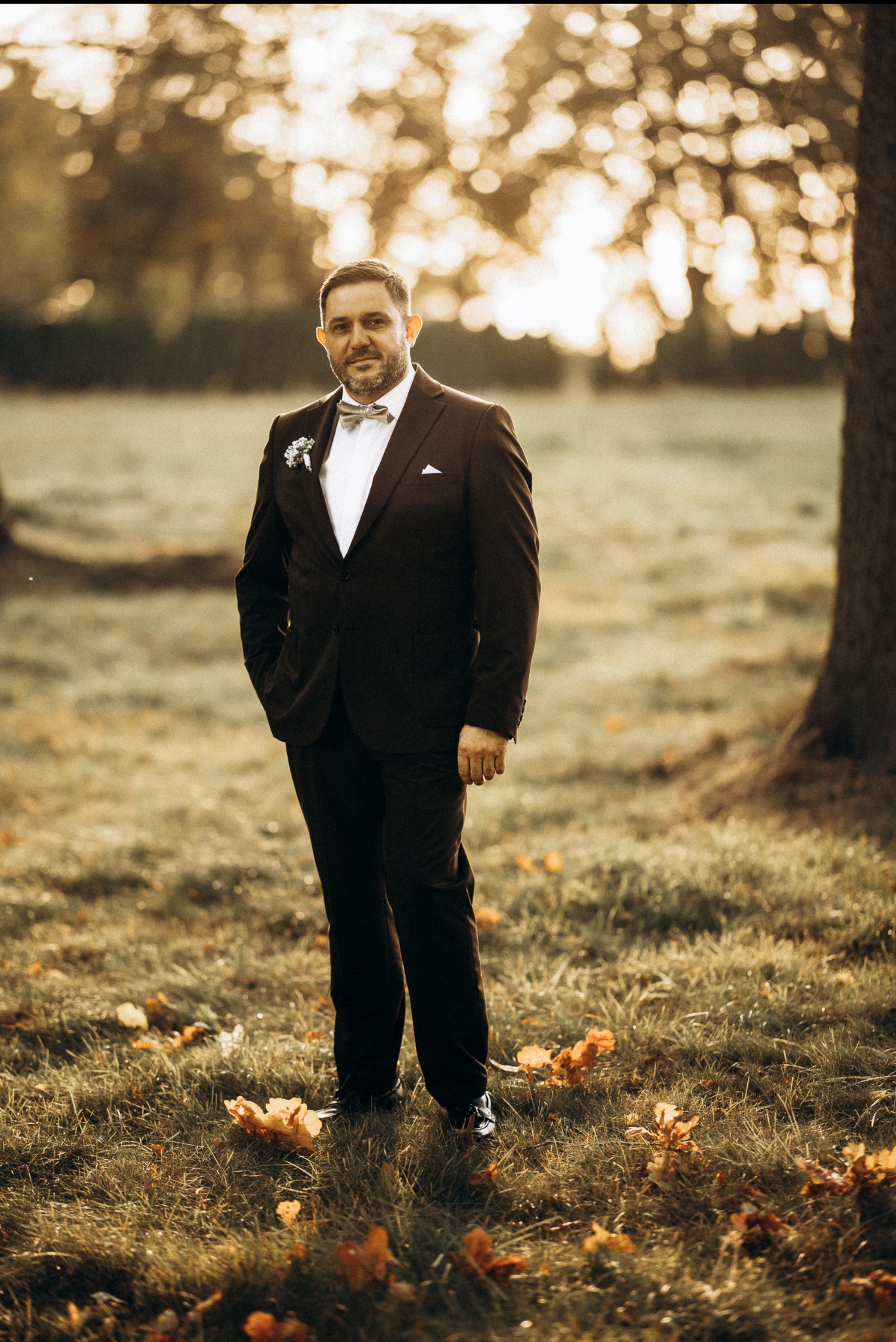 Mężczyzna w brązowym garniturze i muszce, stojący na trawie z opadłymi liśćmi w jesiennej scenerii. Portret w plenerze, ciepłe światło.