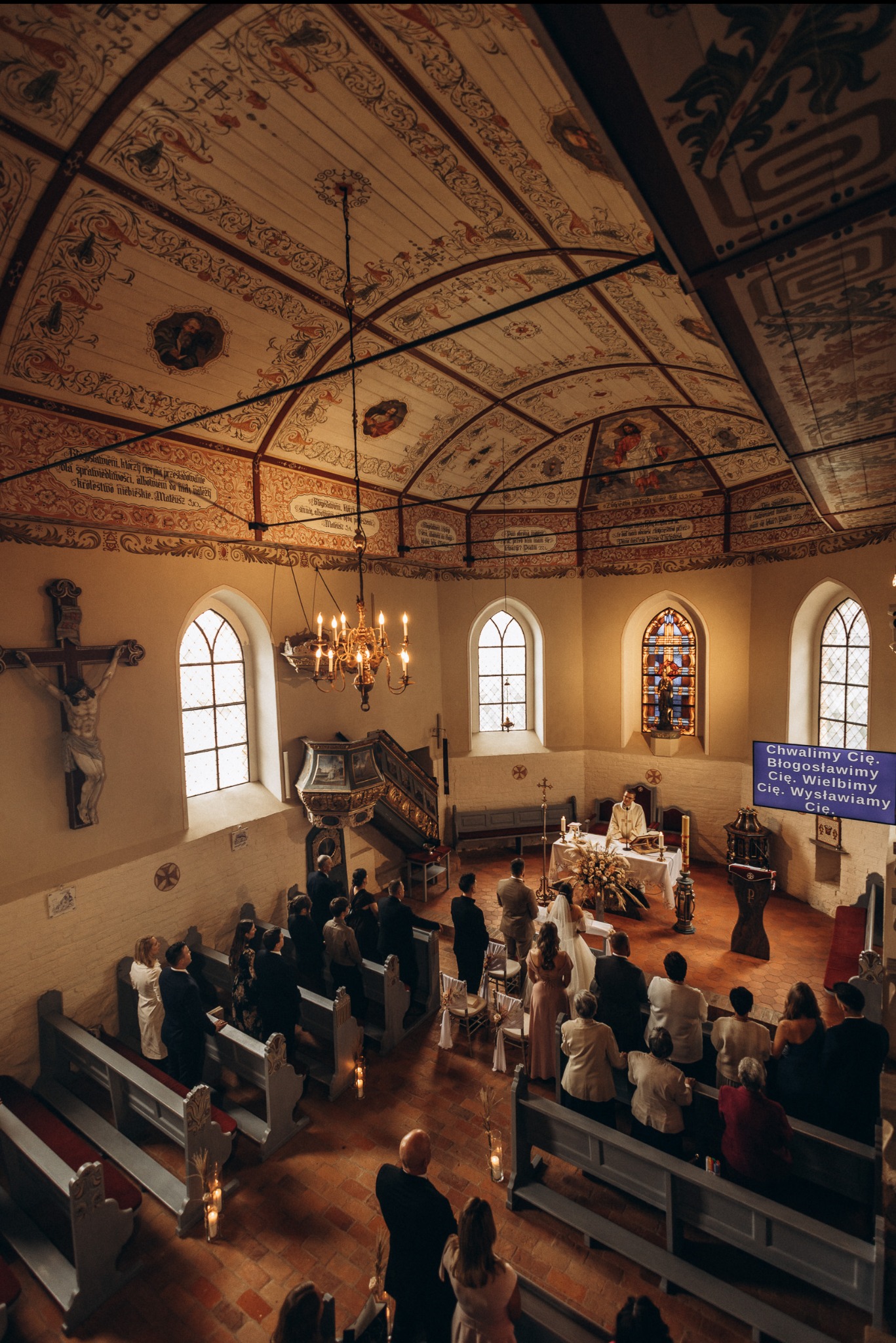 Uroczystość ślubna w kościele z malowanym sufitem, widok z góry na parę młodą przy ołtarzu, gości siedzących w ławkach i księdza odprawiającego mszę.