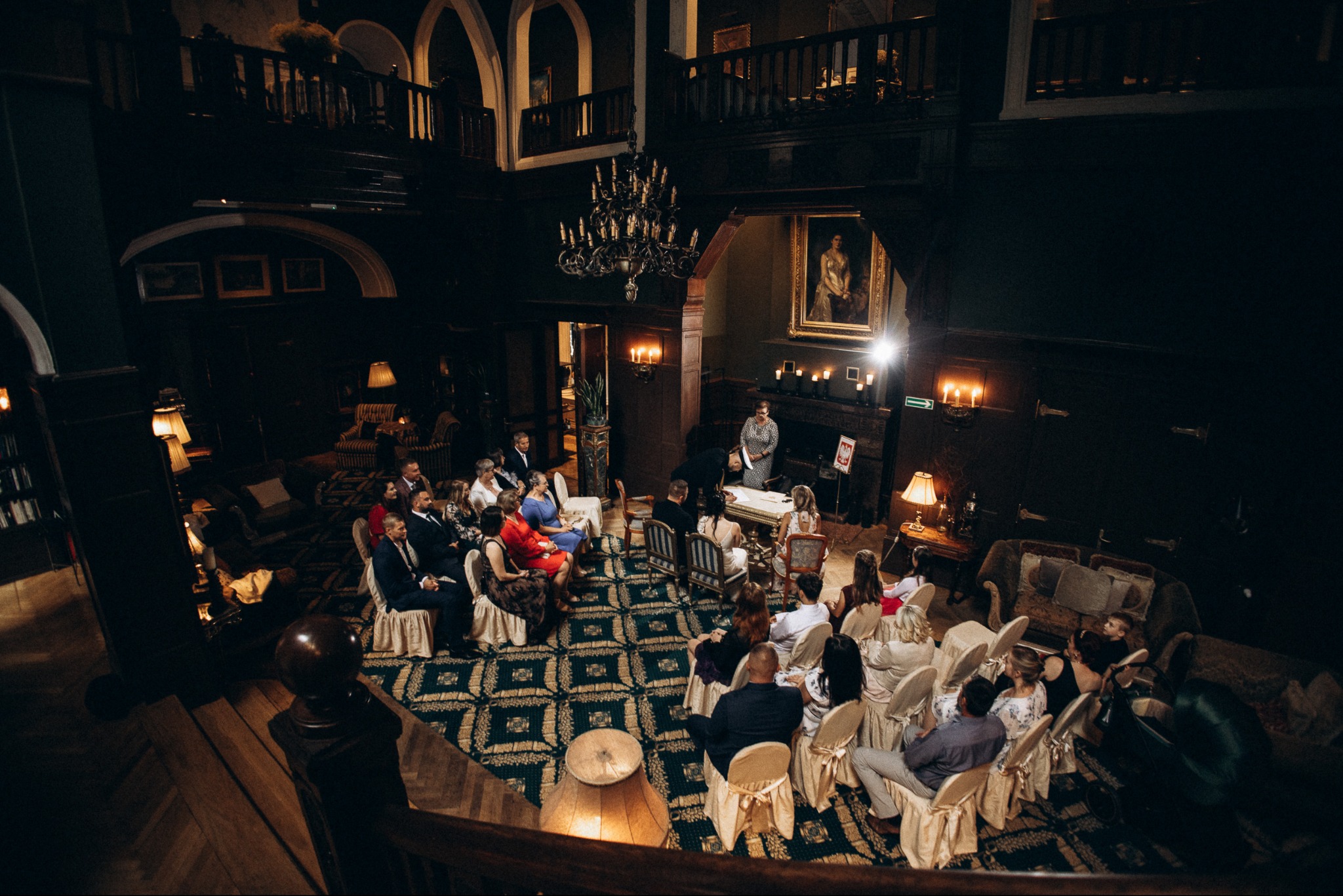 Uroczystość ślubna w Koszalinie: widok z góry na salę z gośćmi, parą młodą przy stole i eleganckim wystrojem wnętrza, z żyrandolem i obrazem.