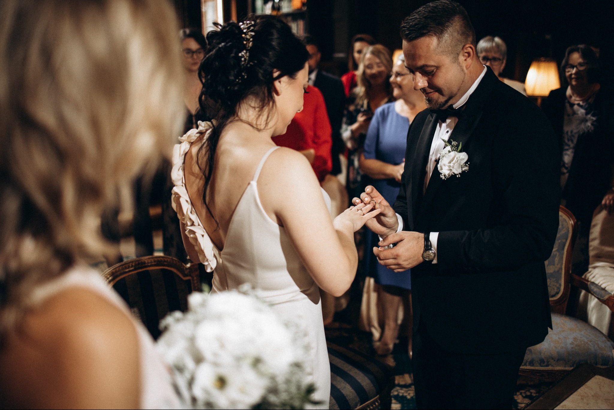 Moment założenia obrączki podczas ceremonii ślubnej. Pan młody w czarnym smokingu, panna młoda w białej sukni. Goście weselni w tle.