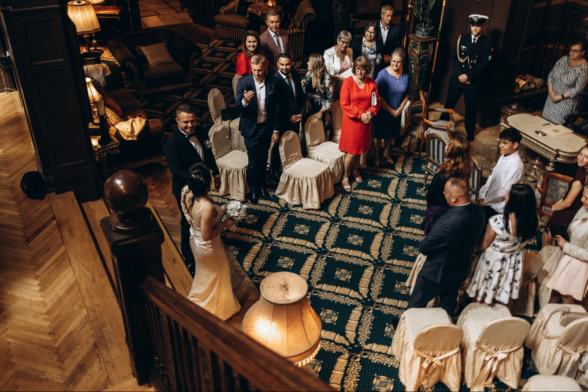 Para młoda na tle gości weselnych w eleganckim wnętrzu. Widok z góry, ukazujący dekoracje, ubiór gości i atmosferę uroczystości. Parkiet w jodełkę i wzorzysty dywan.