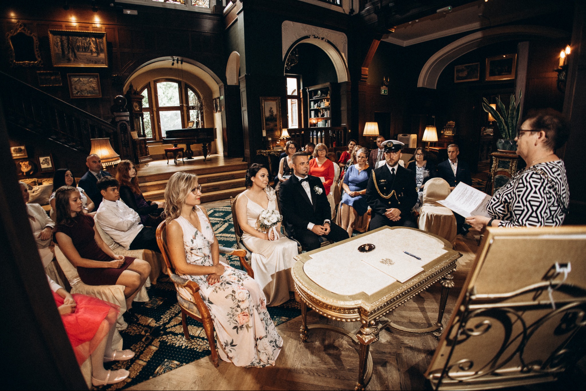 Uroczystość ślubna w stylowym wnętrzu: para młoda siedzi przy zdobionym stole, w tle goście i osoba prowadząca ceremonię. Dekoracyjne oświetlenie i elegancki wystrój.