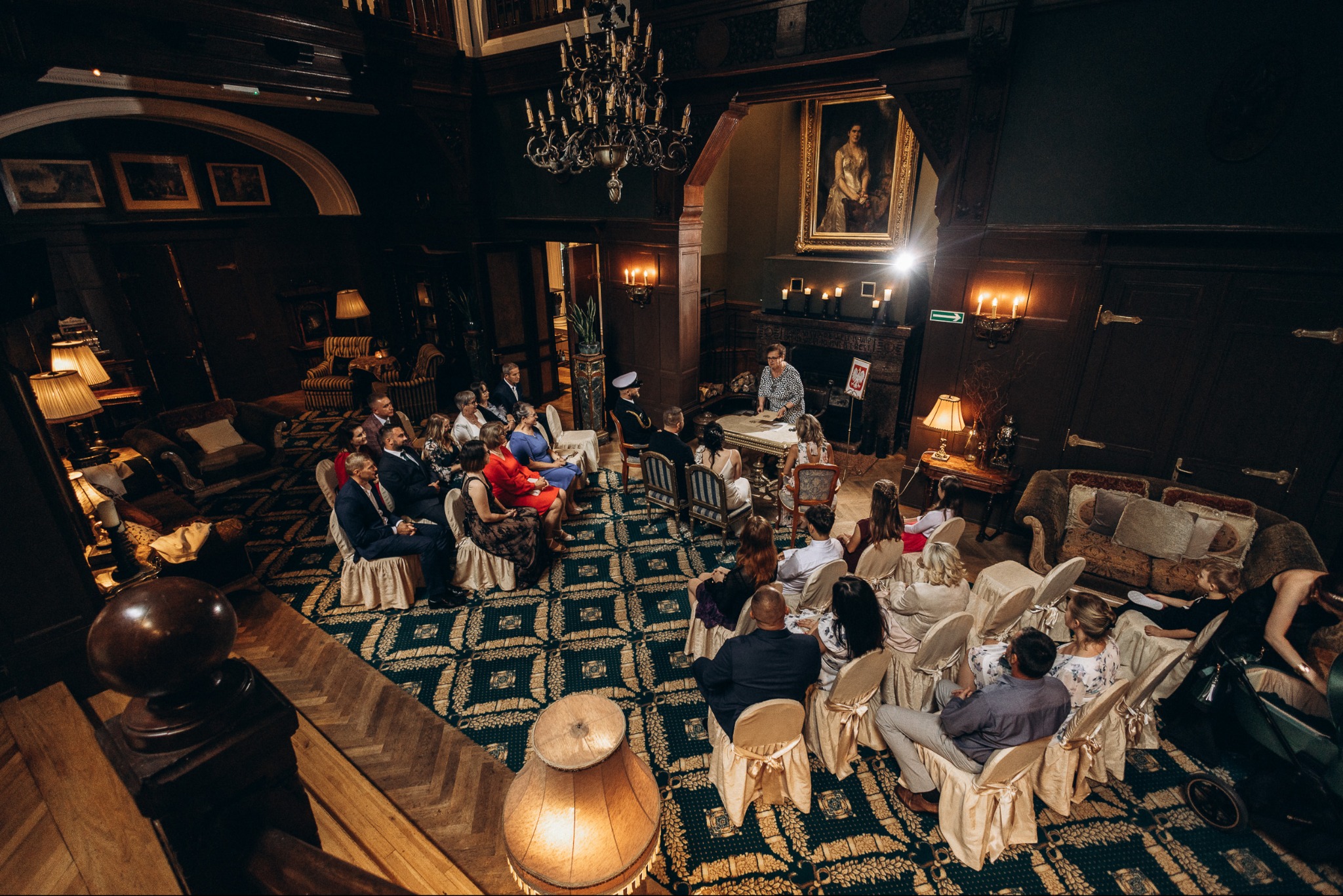 Elegancka ceremonia ślubna w zabytkowym wnętrzu. Goście siedzą na krzesłach z kremowymi pokrowcami, a para młoda stoi przed urzędnikiem na tle kominka.
