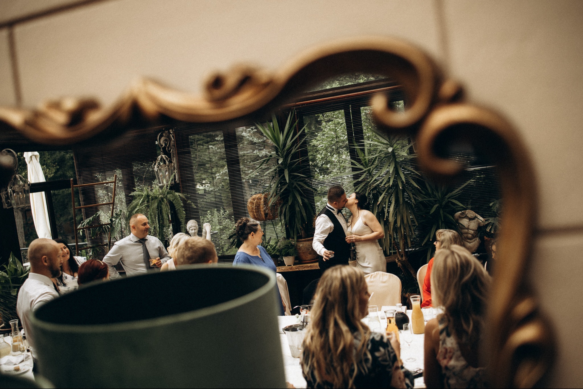 Pocałunek pary młodej uchwycony w ozdobnym lustrze podczas przyjęcia weselnego w oranżerii, z gośćmi w tle. Ciepłe światło.