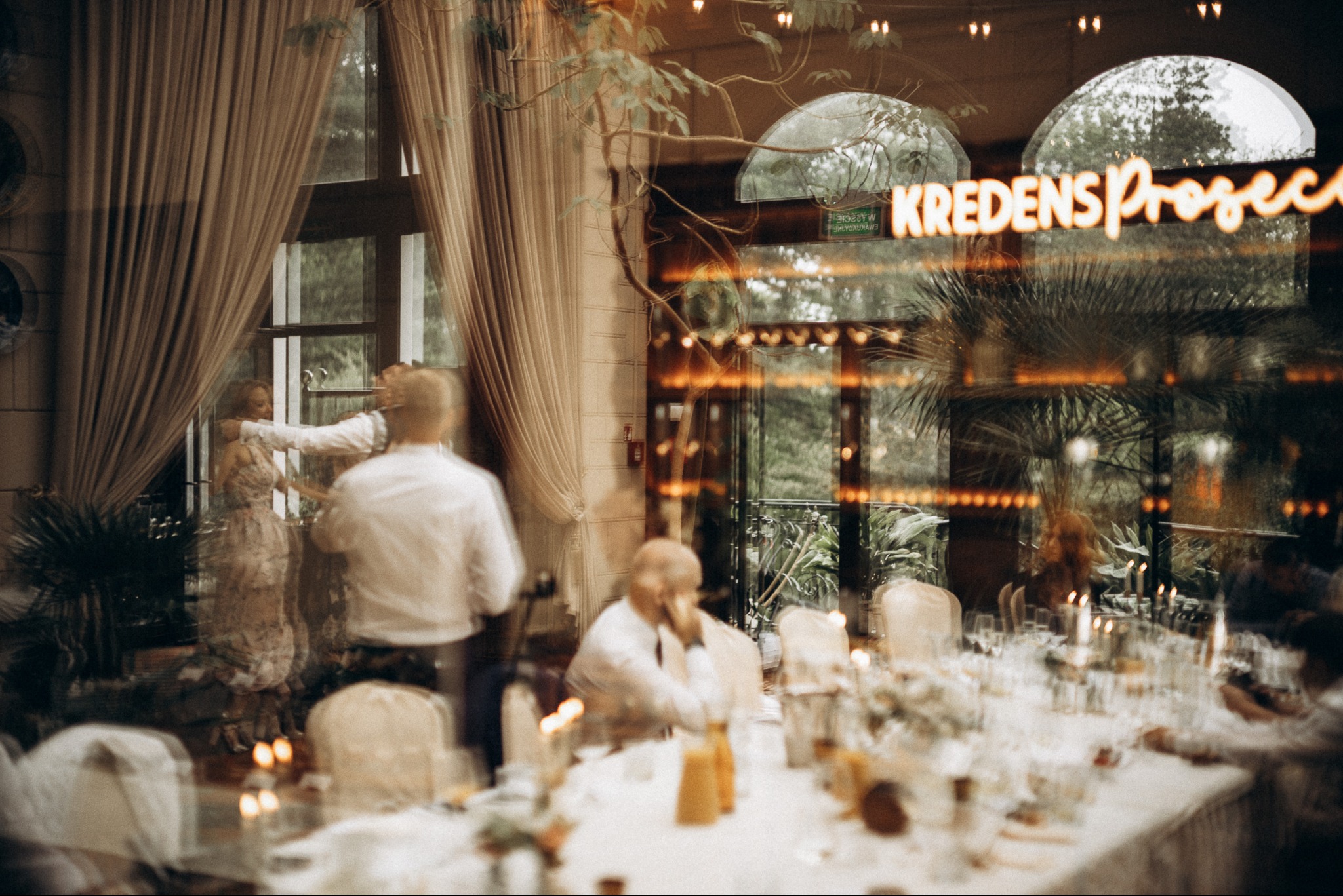 Przygotowania do uroczystości w restauracji: kobieta w sukni poprawiana przez mężczyznę przy oknie, nakryty stół, świece, odbicie neonu 'Kredens Prosecco'.