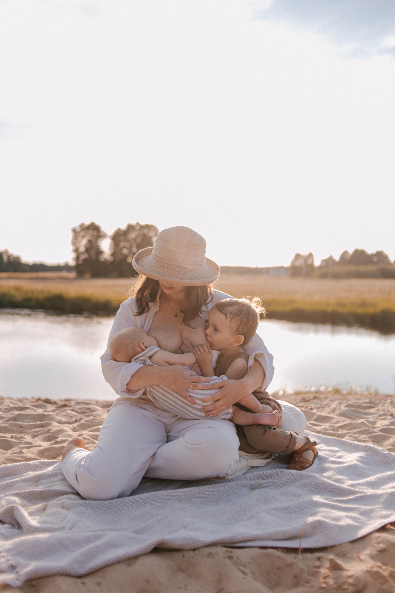 Matka w kapeluszu karmiąca piersią dwoje dzieci na plaży, w tle woda i drzewa.