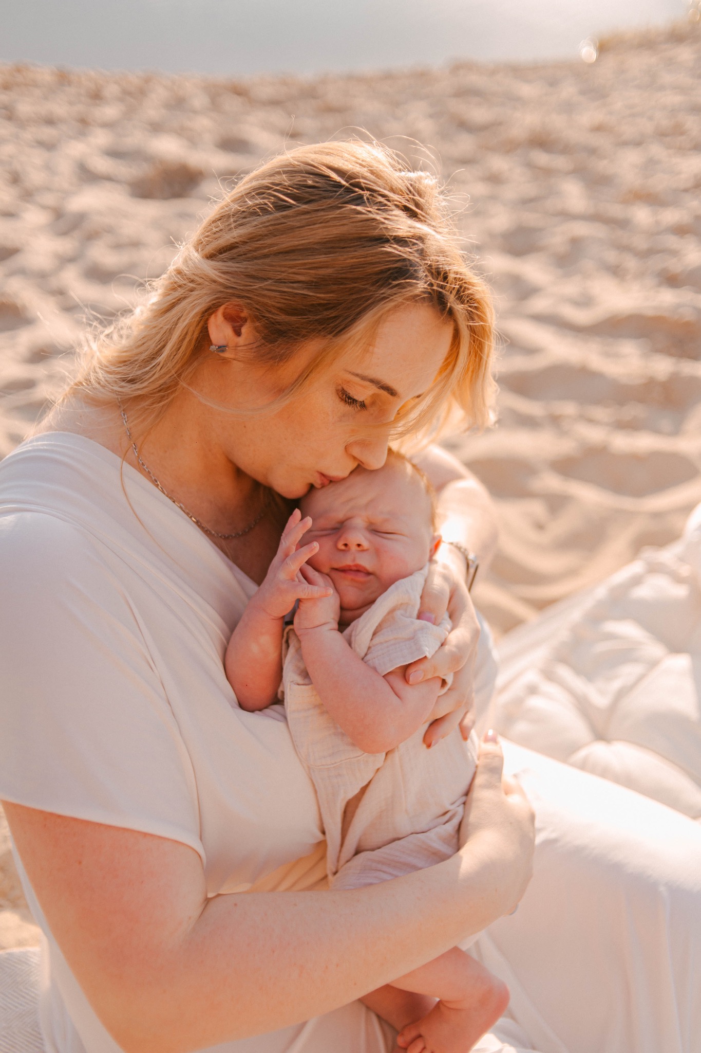 Matka w jasnej sukience całuje śpiącego noworodka w kremowym ubranku na tle piaszczystej plaży w słoneczny dzień.