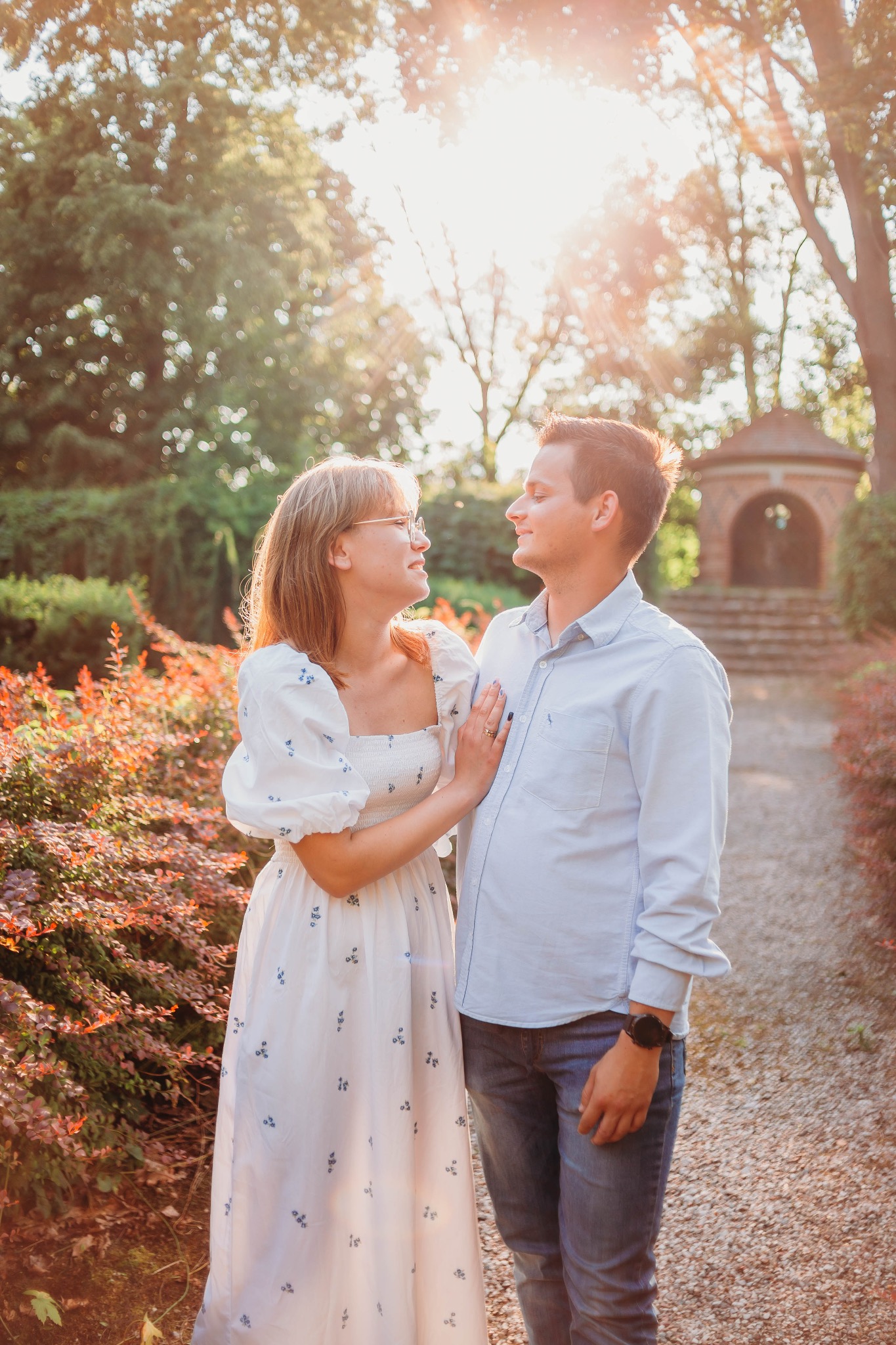 Para zakochanych patrzy na siebie w słonecznym parku, kobieta w białej sukience z niebieskimi kwiatami, mężczyzna w jasnoniebieskiej koszuli i dżinsach, w tle altana.