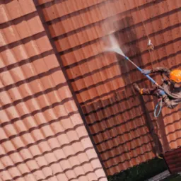 Pracownik w kasku i kombinezonie, zabezpieczony linami, czyści czerwoną dachówkę za pomocą myjki ciśnieniowej. Widoczna różnica w czystości między umytą a brudną częścią dachu, ujęcie z góry.