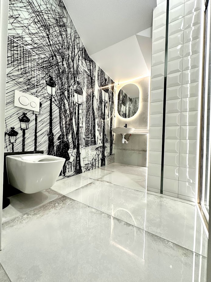 Nowoczesna łazienka z białą ceramiką, błyszczącymi płytkami podłogowymi i ścianą z czarno-białym motywem paryskiej ulicy, oświetlona owalnym lustrem z podświetleniem LED.
