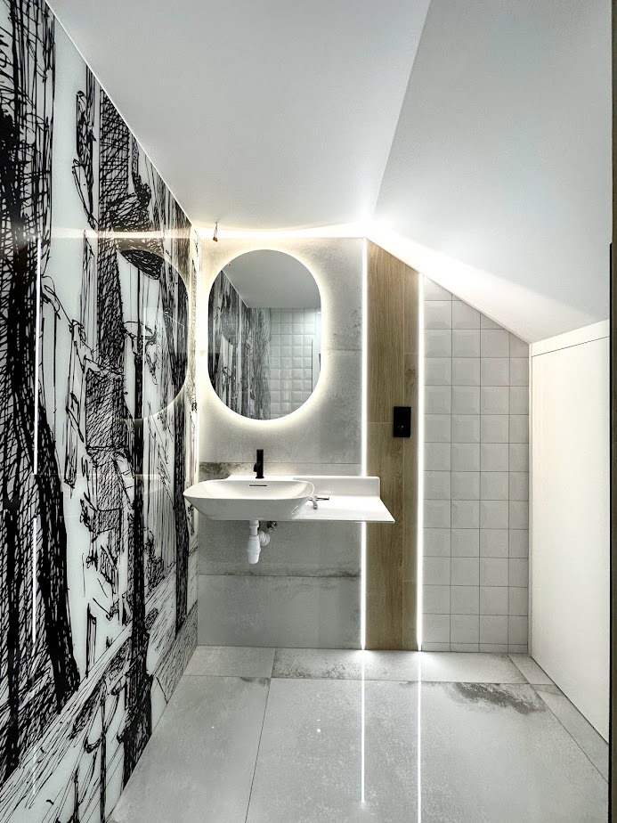 Nowoczesna łazienka na poddaszu z umywalką, okrągłym lustrem z podświetleniem LED, biało-szarą glazurą z motywem urbanistycznym i drewnopodobnymi panelami akcentującymi.