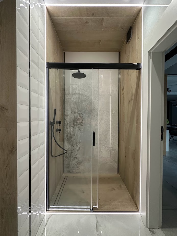 Kabina prysznicowa z przesuwnymi drzwiami, wykończona płytkami imitującymi drewno i białymi, podłużnymi płytkami. Oświetlenie LED wzdłuż krawędzi kabiny i sufitu. Czarna armatura prysznicowa.