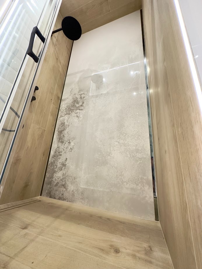 Wykończona kabina prysznicowa z beżowymi płytkami imitującymi drewno na ścianach i podłodze, szklane drzwi z czarnym uchwytem, deszczownica w kolorze czarnym, jasne płytki imitujące beton...