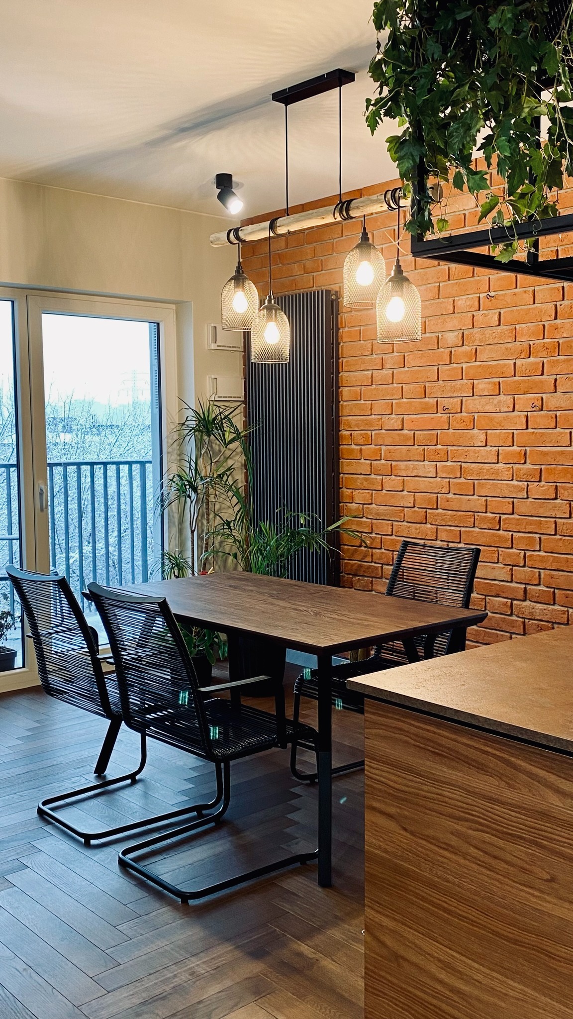 Jadalnia z drewnianym stołem, plecionymi krzesłami, ceglaną ścianą i wiszącą lampą z żarówkami w drucianych kloszach, podkreślona zielenią roślin w doniczkach.