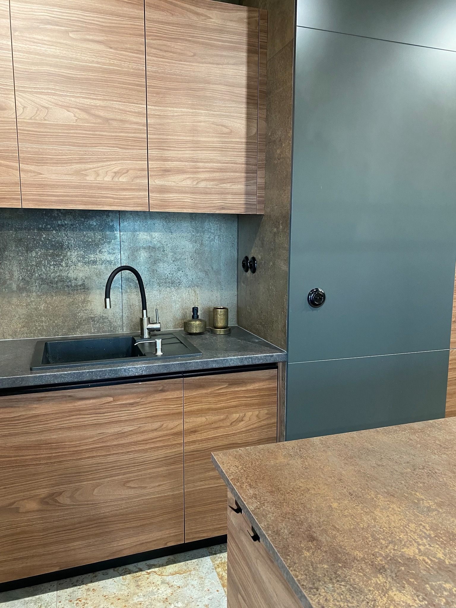 Nowoczesna kuchnia z drewnianymi frontami szafek, czarnym zlewem i baterią, oraz szarą zabudową sięgającą sufitu. Blat z ciemnego kamienia i dekoracyjne pojemniki obok zlewu.