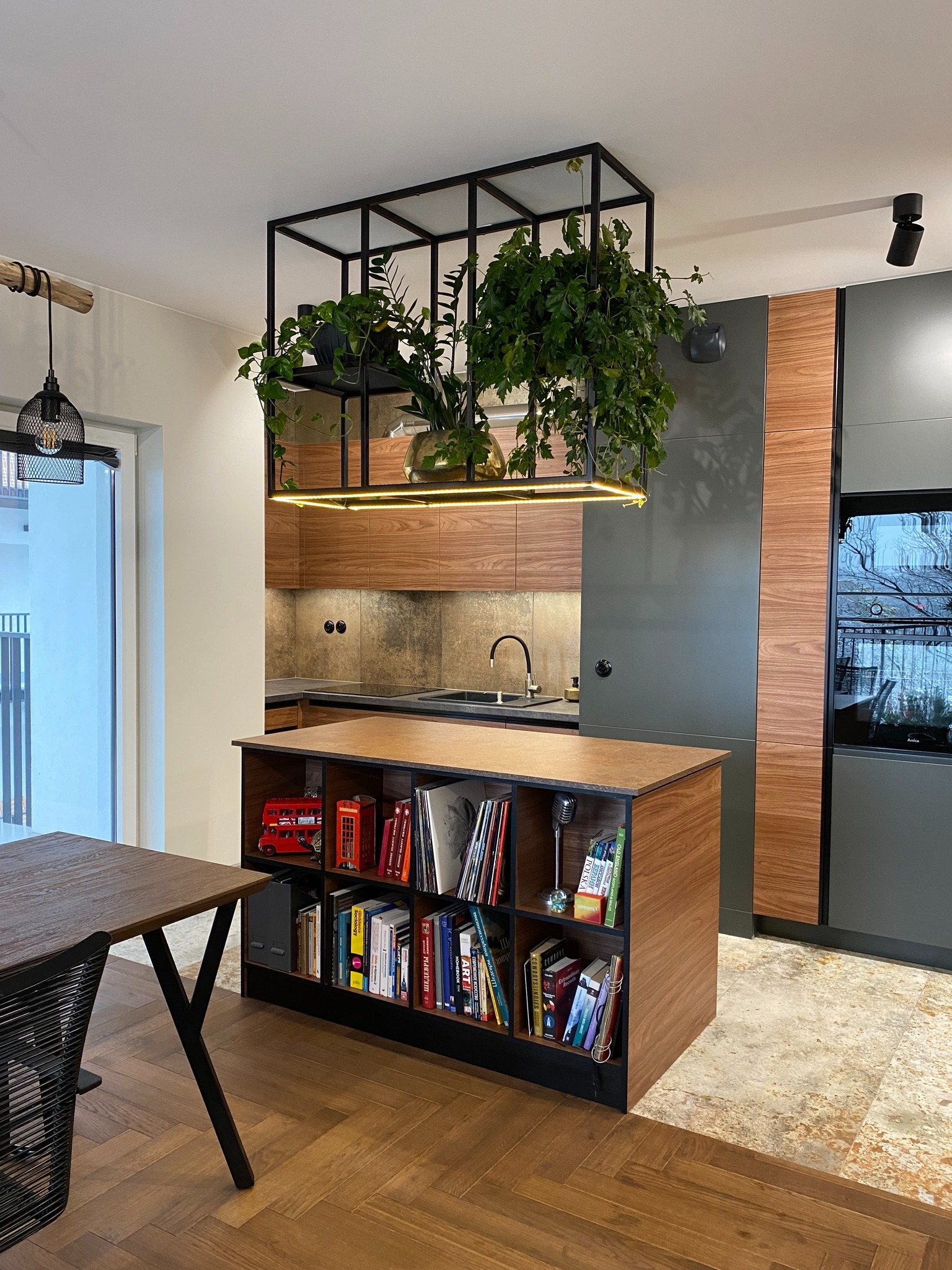 Nowoczesna kuchnia z wyspą i wiszącą konstrukcją z roślinami, książkami i dekoracjami. Na wyspie widoczny czerwony autobus i budka telefoniczna jako ozdoby.