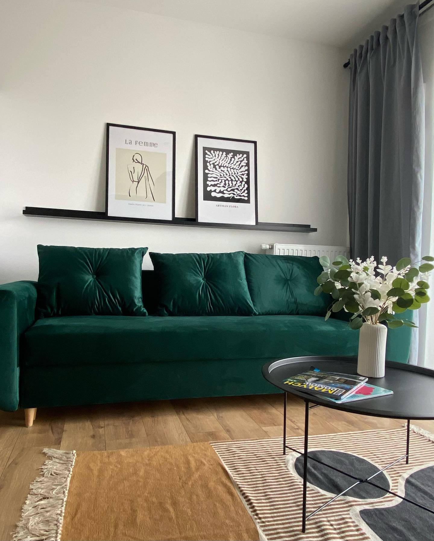 Salon z zieloną sofą z weluru, dekoracyjnymi poduszkami, czarną półką z plakatami, okrągłym stolikiem kawowym i dywanami w różnych odcieniach brązu.