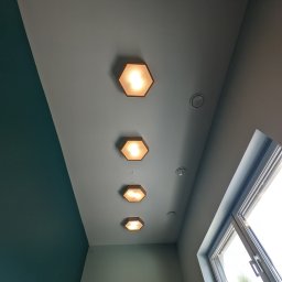 Oleksandr Mazur - Pomieszczenie po remoncie: gładki, szary sufit z rzędem czterech sześciokątnych lamp z ciepłym światłem, widoczne nawiewniki, ściana pomalowana na dwa kolory (szary i ciemnozielony), okno...
