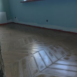 Oleksandr Mazur - Ułożona pod kątem, drewnopodobna podłoga z płytek w jasnym pomieszczeniu z błękitnymi ścianami, widoczne niedoskonałości na styku ze ścianą i oknem, sugerujące prace w toku.