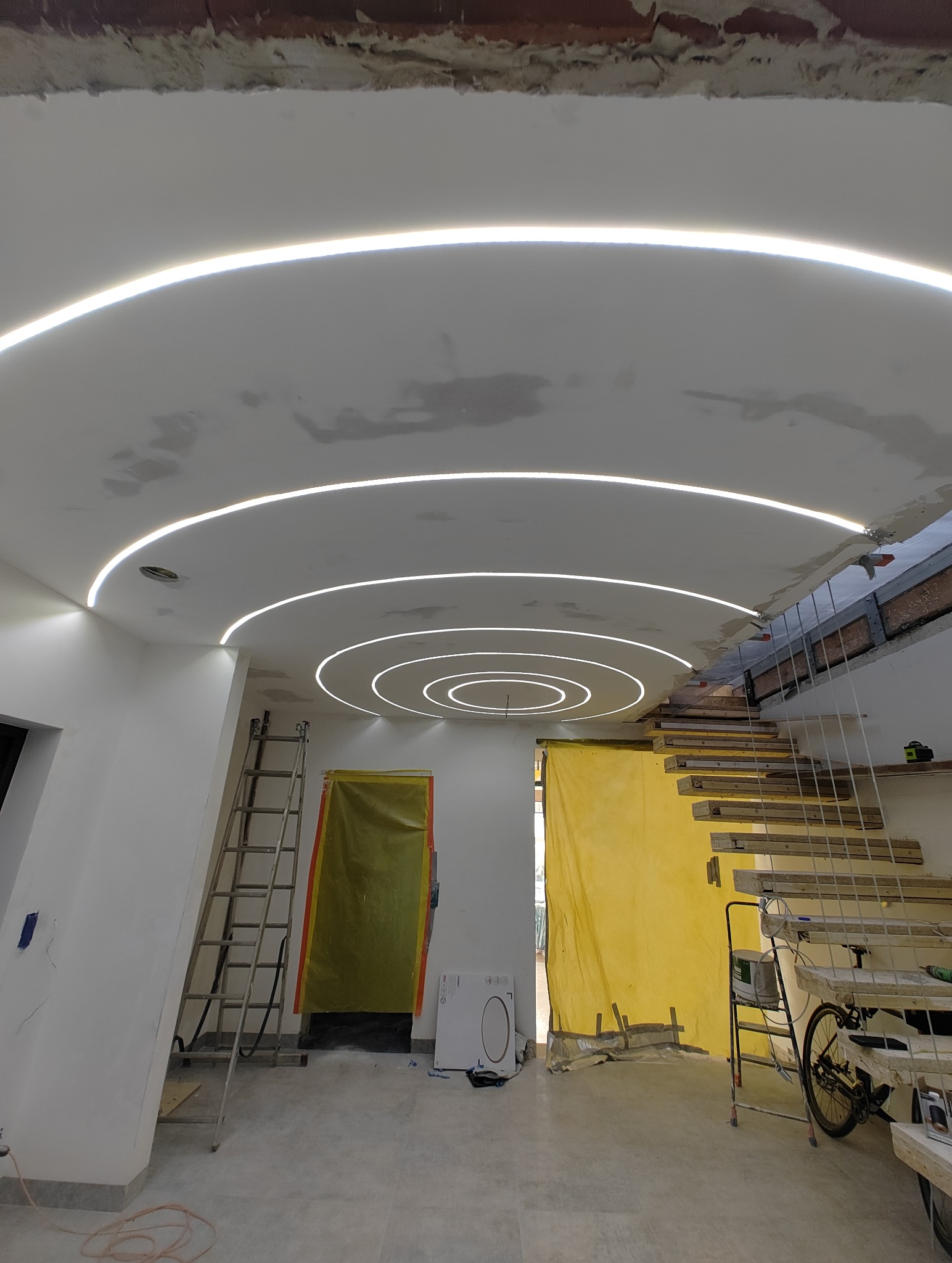 Instalacja sufitu podwieszanego z oświetleniem LED w kształcie koncentrycznych okręgów, widoczne drabiny i zabezpieczenia folią malarską, surowy stan wykończenia.