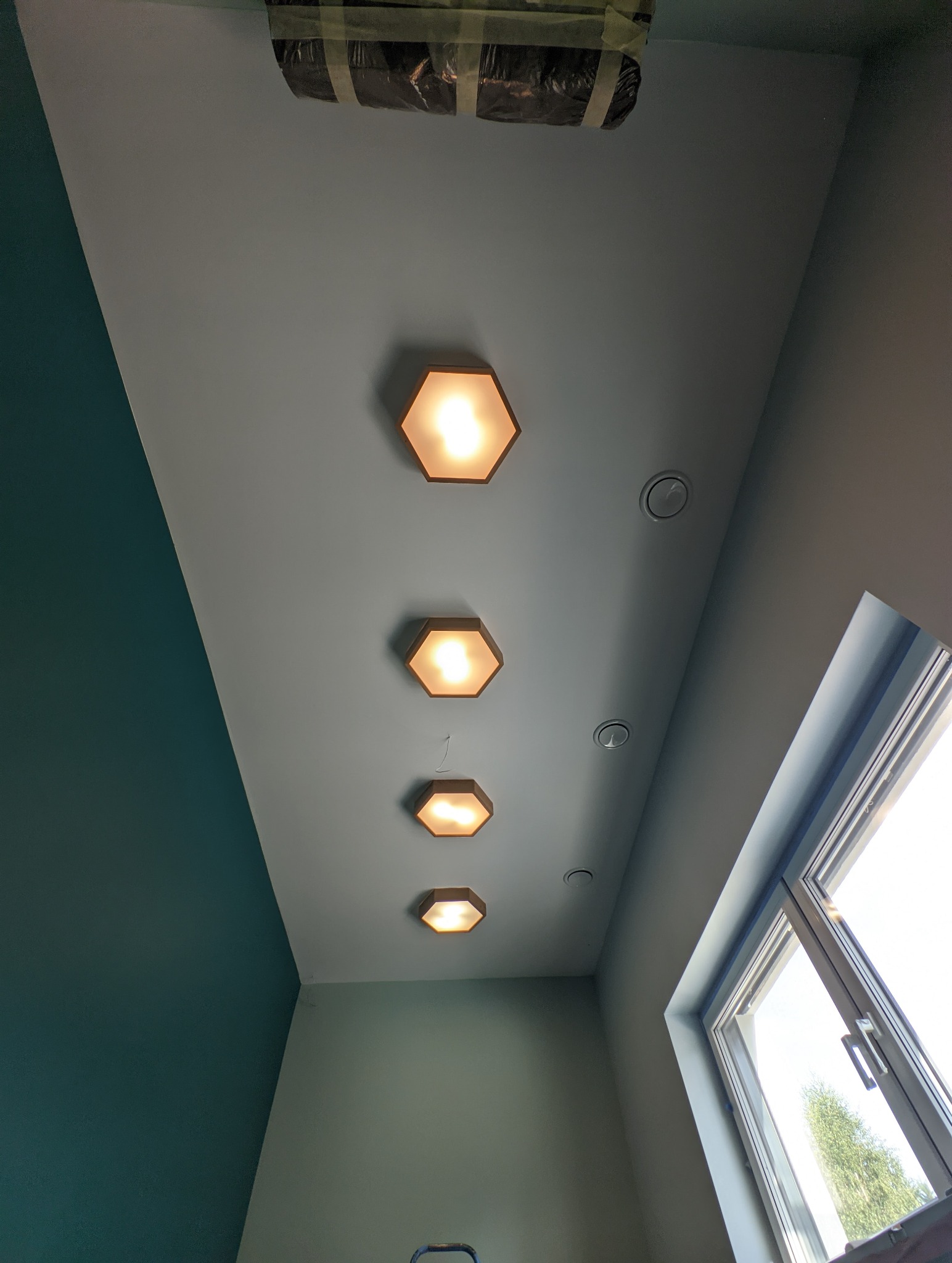 Pomieszczenie po remoncie: gładki, szary sufit z rzędem czterech sześciokątnych lamp z ciepłym światłem, widoczne nawiewniki, ściana pomalowana na dwa kolory (szary i ciemnozielony), okno...