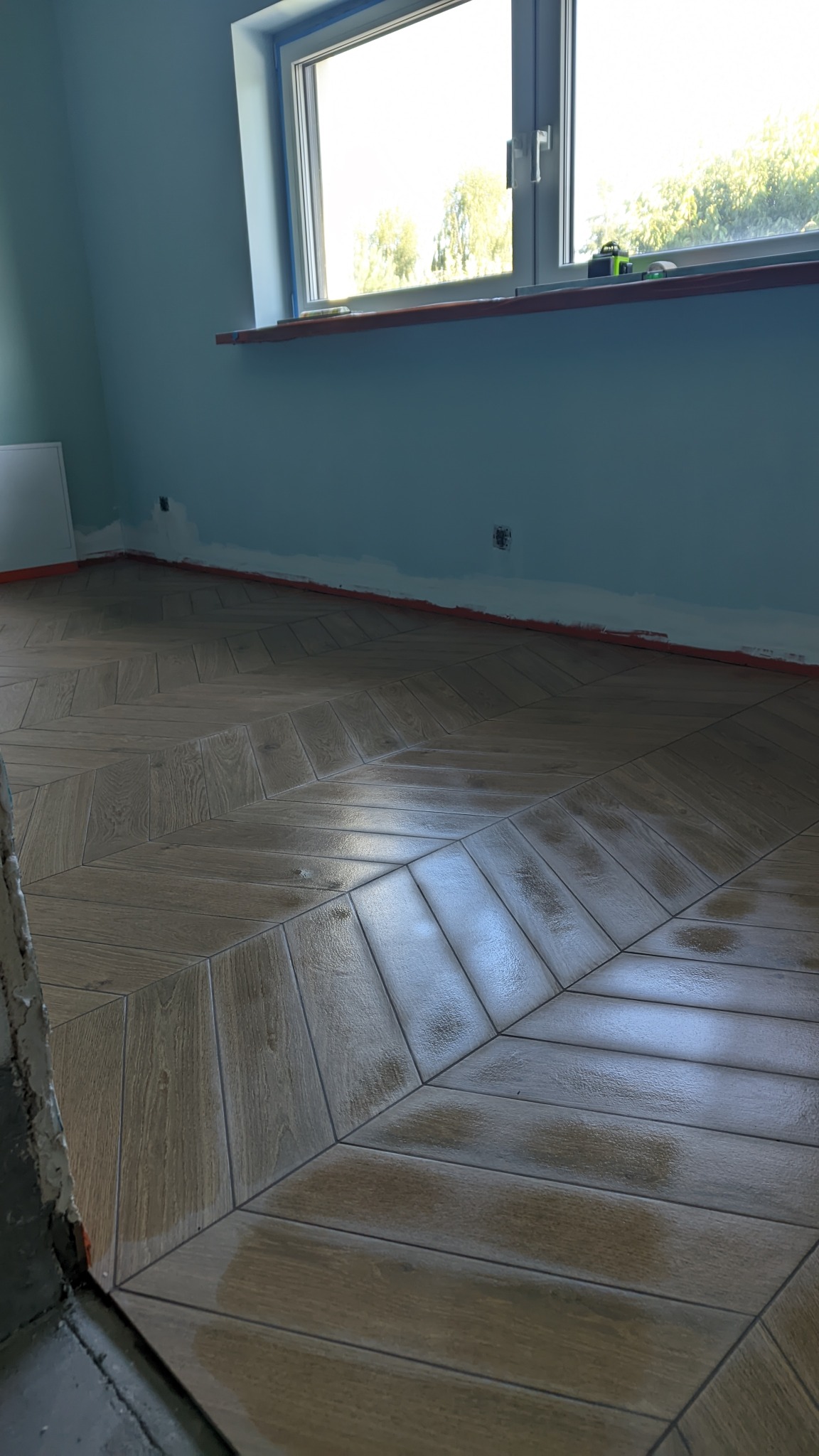 Ułożona pod kątem, drewnopodobna podłoga z płytek w jasnym pomieszczeniu z błękitnymi ścianami, widoczne niedoskonałości na styku ze ścianą i oknem, sugerujące prace w toku.