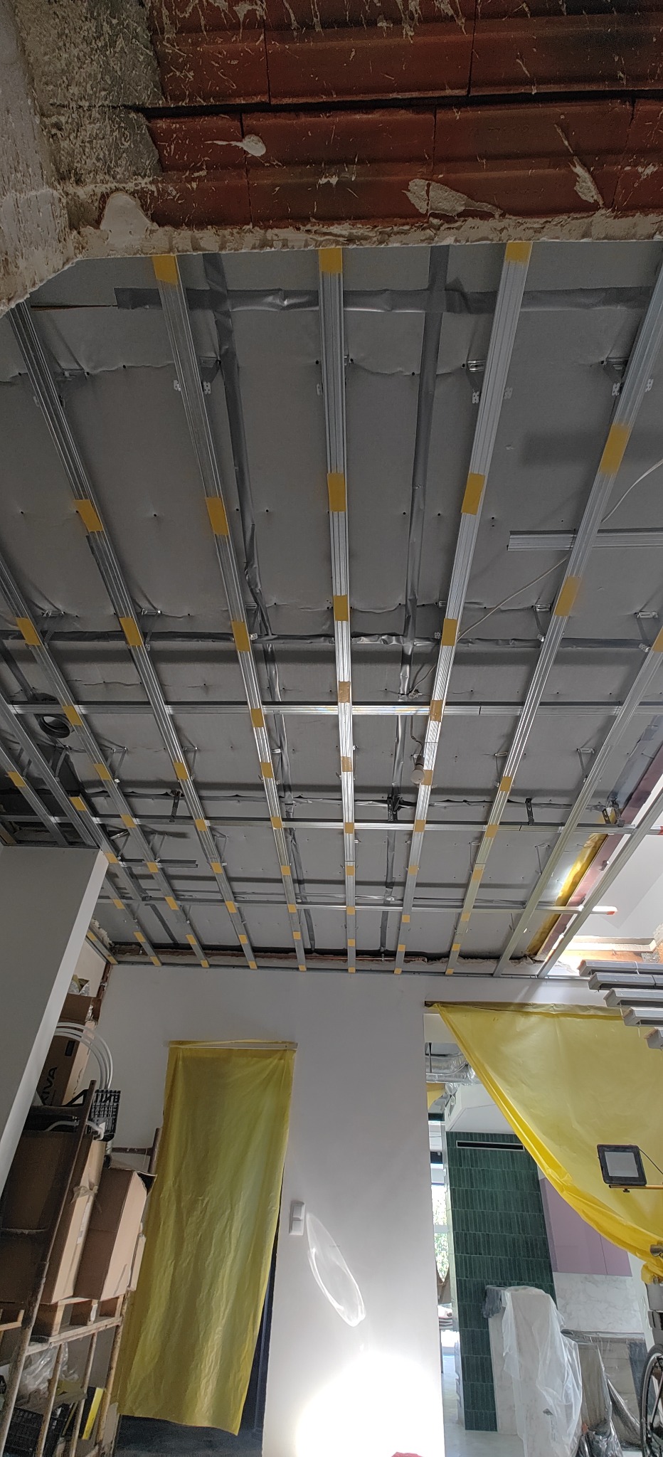 Stelaż sufitu podwieszanego z płyt kartonowo-gipsowych, widoczne profile metalowe z żółtymi taśmami mocującymi, surowy mur w tle, pomieszczenie w trakcie remontu, osłonięte folią malarską.