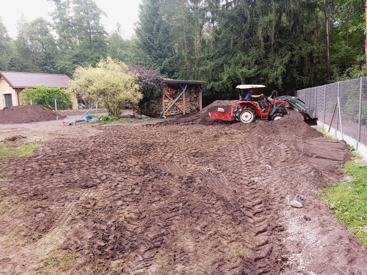 Czerwony traktor wyrównuje teren pod nowy ogród. Widoczne ślady opon na świeżo przekopanej ziemi, w tle budynek i drzewa.