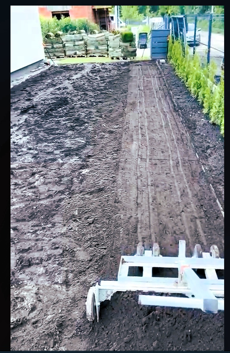 Przygotowanie gleby pod trawnik: wyrównana ziemia z użyciem glebogryzarki, w tle palety z rolkami trawy i tuje. Widok z perspektywy operatora maszyny.