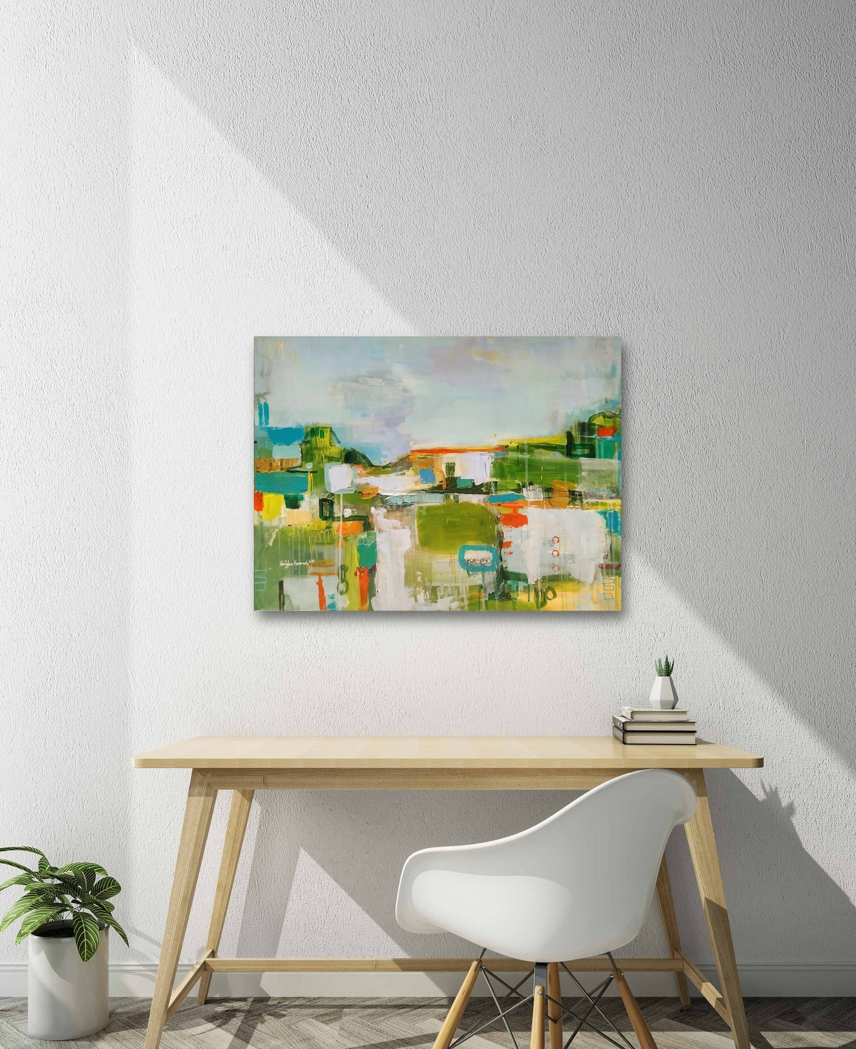 Abstrakcyjny obraz z przewagą zieleni i błękitu zawieszony na białej ścianie nad biurkiem z jasnego drewna, obok białe krzesło i doniczka z rośliną.