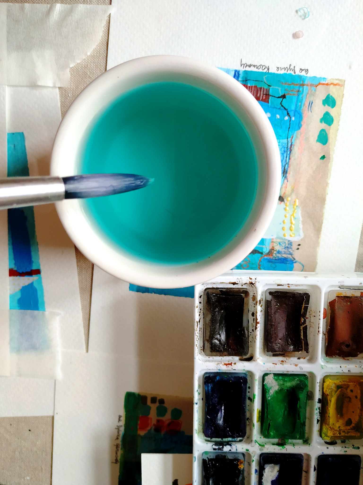Paleta farb akwarelowych, pędzel zanurzony w kubku z turkusową wodą, fragmenty prac malarskich na papierze w tle.
