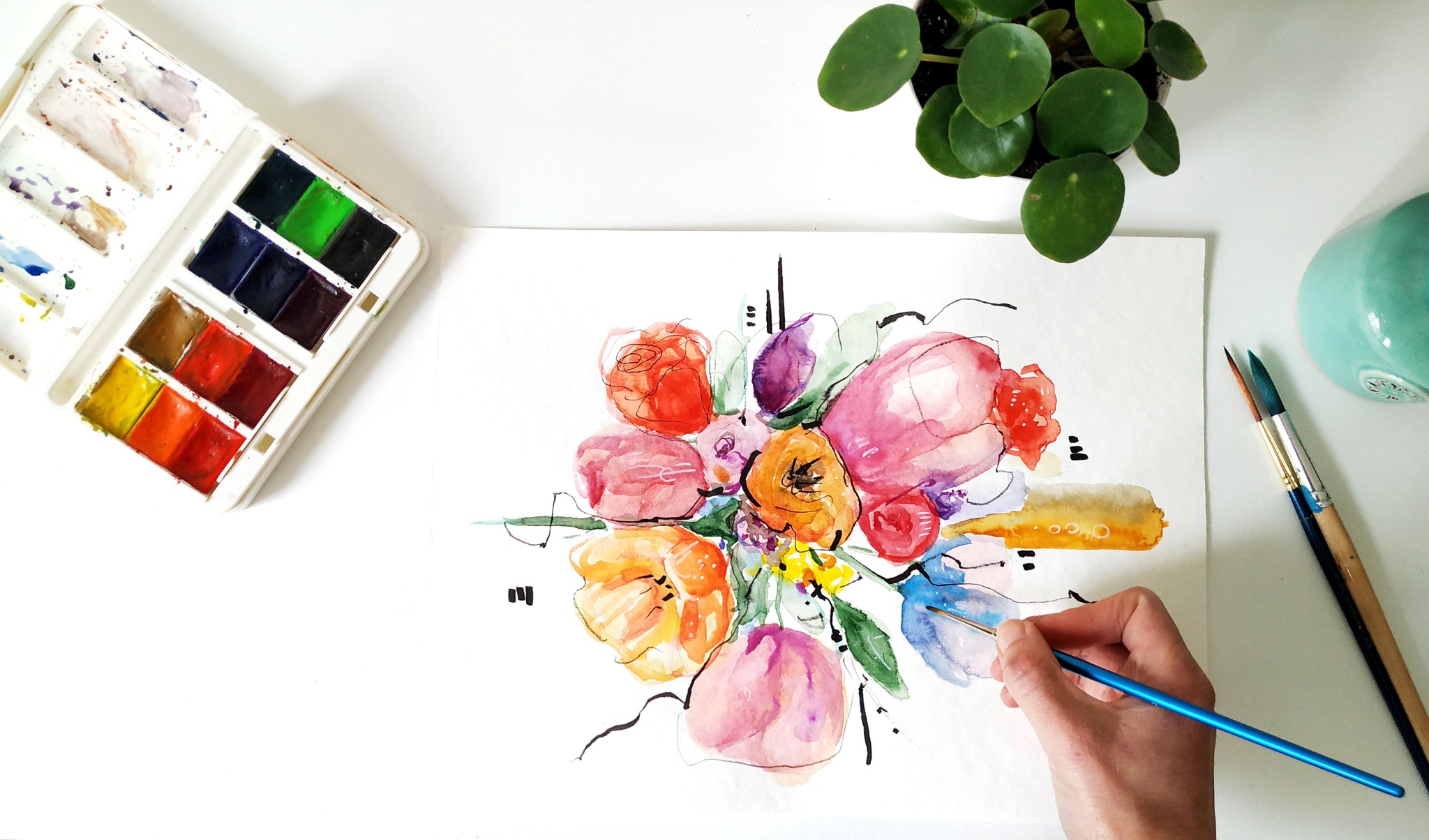 Artystyczne ujęcie z góry: ręka malująca akwarelami bukiet kwiatów na białej kartce, obok paleta z farbami, pędzelki i mała roślina doniczkowa.