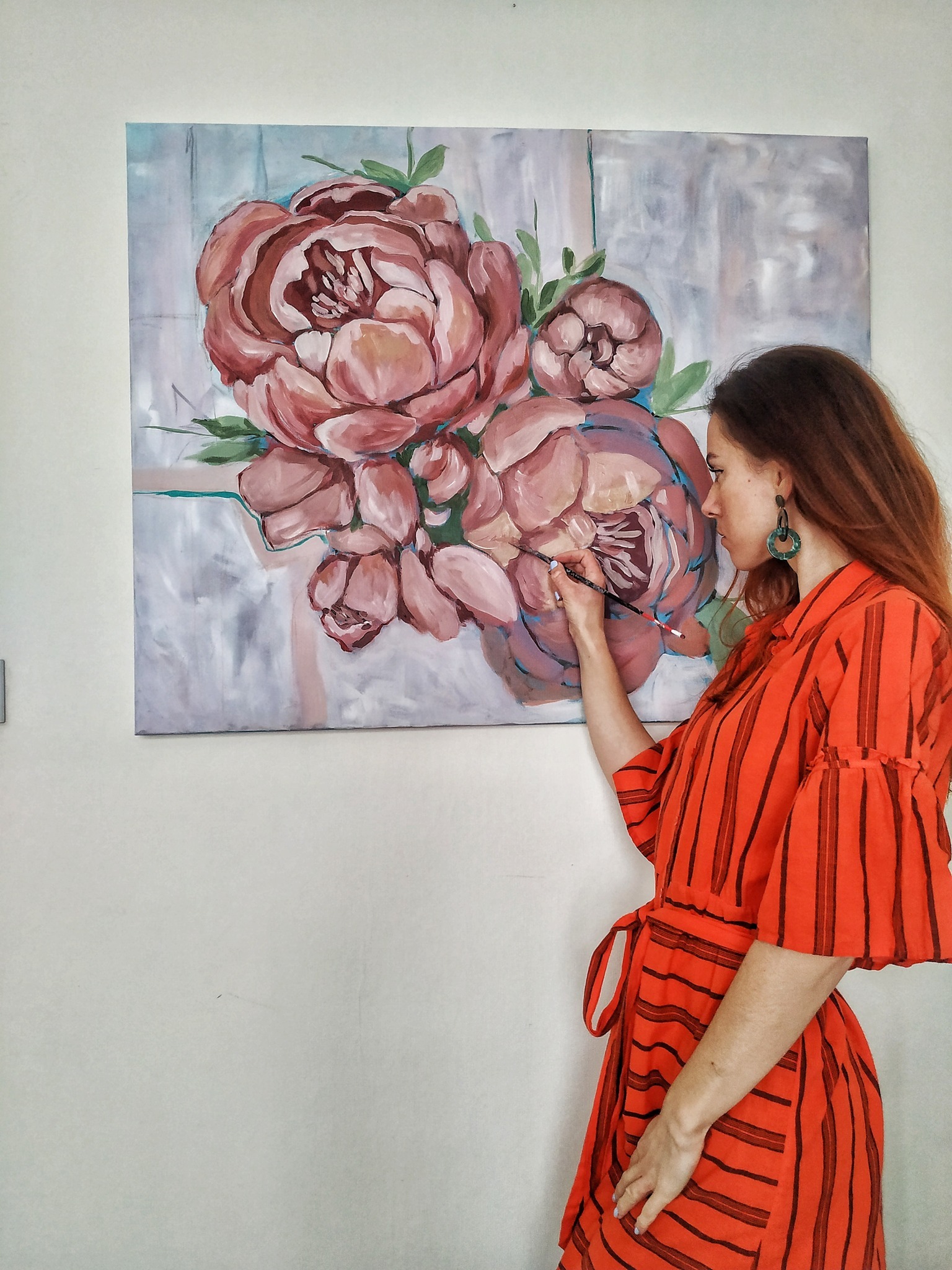 Artystka w pomarańczowej sukience maluje pędzlem obraz z motywem różowych piwonii na płótnie.