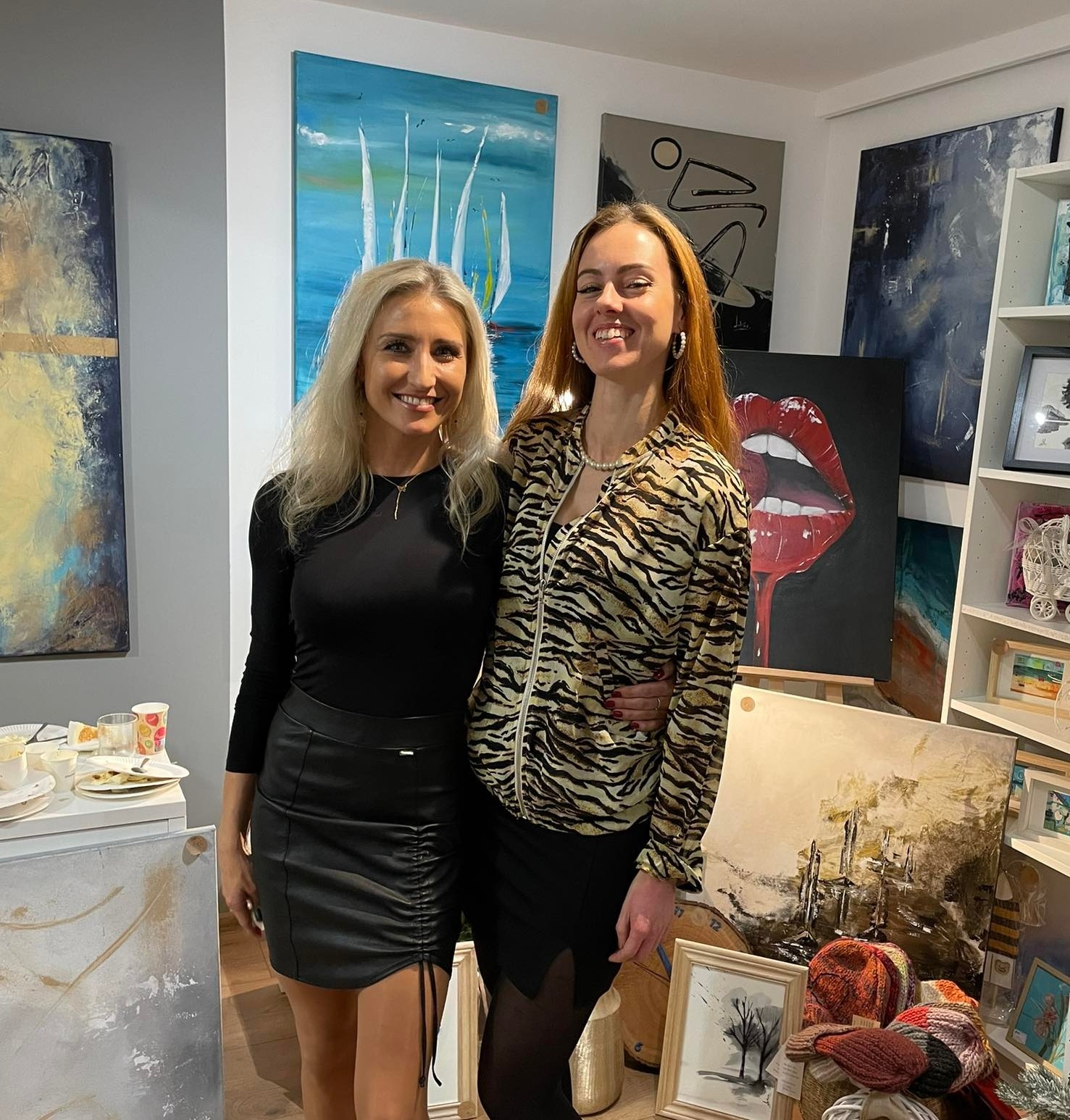 Dwie uśmiechnięte kobiety pozują w galerii sztuki, otoczone obrazami o różnej tematyce, w tym pejzaże morskie i abstrakcje, z widocznymi śladami po poczęstunku na stole.