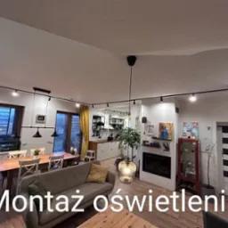 Salon z zamontowanym systemem szynowym oświetlenia punktowego, oświetlającym stół jadalny i przestrzeń wypoczynkową z kominkiem.